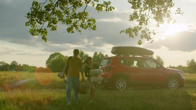 Young Diverse Traveling Cheerful Couple with Blanket and Ukulele goes from suv Car to Picnic on Sunset. Rental Car concept. Steadicam gimbal follow wide shot in slowmotion. Shot on RED Cinema camera