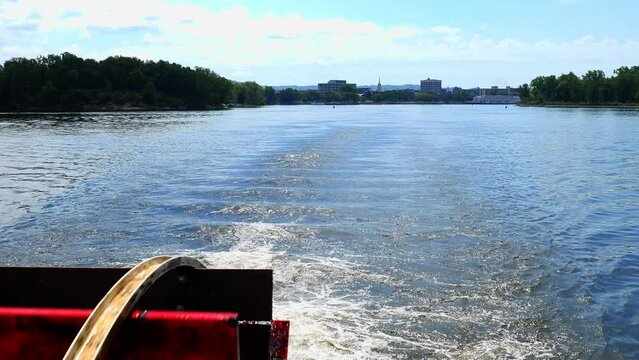 Red Wooden Paddle-wheel turning and causing foamy Mississippi River water, with La Crosse, Wisconsin in the distance.