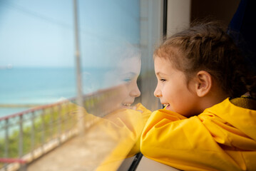 A little girl of 6-7 years old looks out the window of a train at the sea. She's wearing a yellow parka. Journey. Reflection. Vacation. Summer. Family vacation.