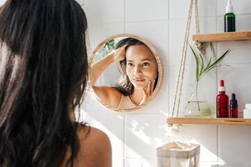 Beautiful hispanic woman looking in the mirror in white eco friendly bathroom. Wooden shleves and reusable cosmetics bottles. Wellnes concept - obrazy, fototapety, plakaty