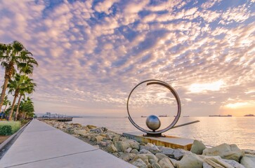 Molos Promenade and skyline of the coast in Limassol city in Cyprus at cloudy sunrise. View of boardwalk pier path landmark with palm trees, sculpture, the Mediterranean sea and people walking.	
