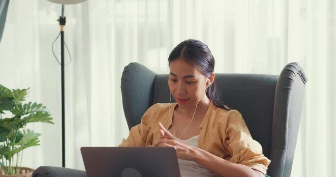Young asia female girl or university student sit on sofa chair with computer laptop wear earphone listen music chill and singing take a rest work in living room at house. Home lifestyle concept.