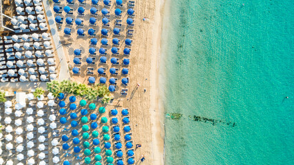 Aerial above bird's eye view of Pantachou - Limanaki organised beach (Kaliva), Ayia Napa, Famagusta, Cyprus. Blue aligned umbrellas, golden sand, parasols, sunbathing sea beds clean turquoise water	
