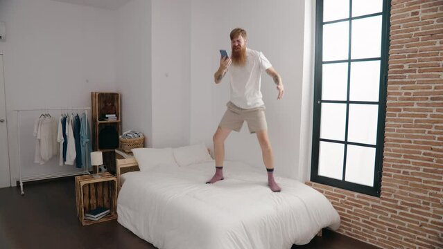 Young redhead man singing song using smartphone standing on bed at bedroom