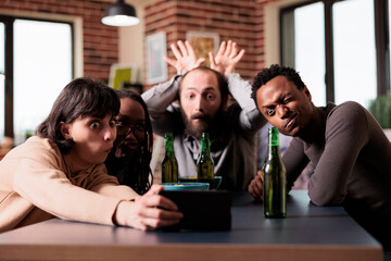Goofy childish multicultural group of friends with smartphone device taking selfie photo. Playful diverse people sitting at home in living room while taking photo together.