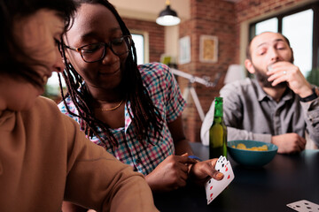 Smiling african american woman showing cards to friend while sitting in living room. Happy diverse people sitting at home while enjoying fun leisure activity together with snacks and beverages.