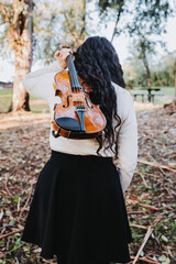 curly brunette woman holding a violin on her back, walking through the park road at sunset. Vertical