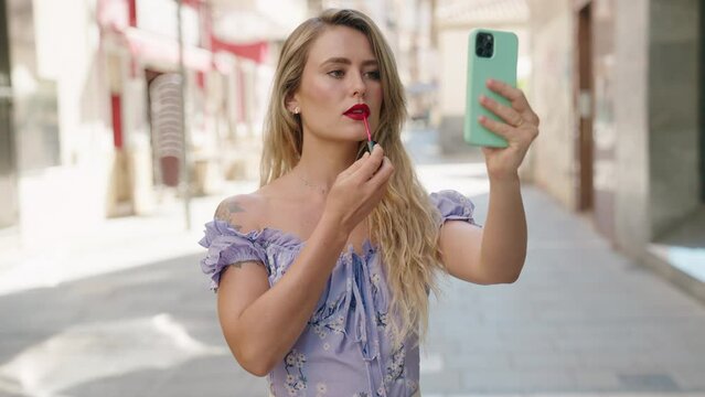 Young beautiful hispanic woman using smartphone as a mirror make up lips at street