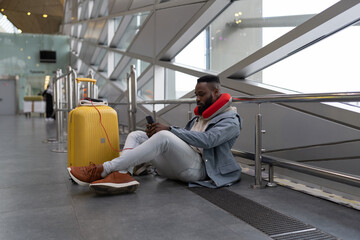 Fototapeta premium Pensive attractive African American guy blogger using mobile phone, communicating in social media, writing post about trip, searching information online while sitting in airport or railway station. 