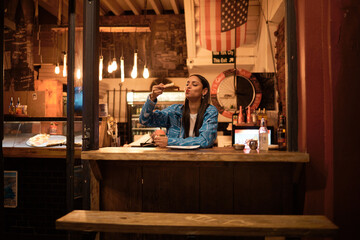 A young and happy woman eating pizza sitting alone in an empty and dark American restaurant. A...