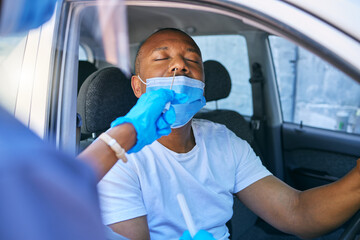 Covid testing and screening of a man driving in his car at a drive through station with medical nurse assistance. Guy getting virus treatment test while wearing a protective face mask