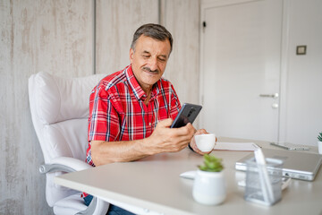 senior man hold cup of coffee and mobile phone while sitting at work