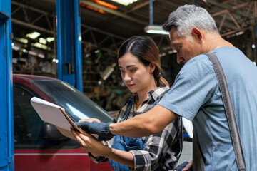 Senior Asian mechanic teaches a female trainee holding a clipboard to write down the list of engines that need to be repaired in the garage. small business and car repair service concept