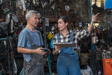 Fototapeta na wymiar Senior Asian mechanic teaches a female trainee holding a clipboard to write down the list of engines that need to be repaired in the garage. small business and car repair service concept