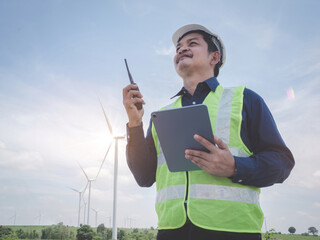 Asian engineer man stand holding tablet front the wind turbines generating electricity power station with talking by walkie -talkie . Concept of sustainability development by alternative energy