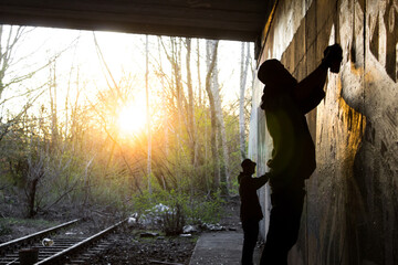 graffiti artist painting under a bridge while sunset on a wall - dark silhouette of the artists in dark shadow