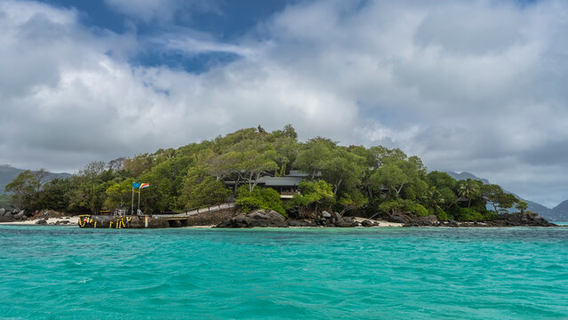 A small secluded tropical island overgrown with lush green vegetation. The house is visible through the branches. Pier in the aquamarine ocean. Clouds in the blue sky. Seychelles