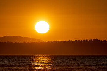 A brilliant orange sun sets over Puget Sound with silhouetted hills in the background and reflections on the ocean in the foreground