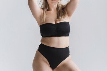 young overweight woman in sports minimalist black lingerie poses  near large window. Plus size model show body. Folds at the waist.  Unrecognizable plus size faceless model