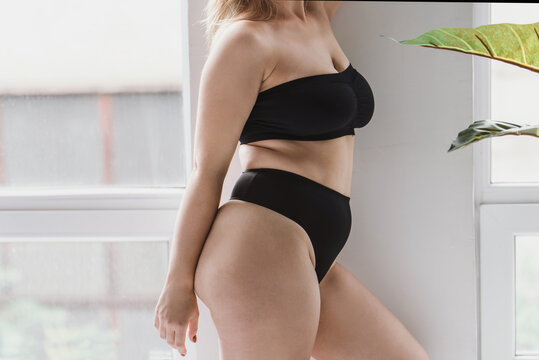 young overweight woman in sports minimalist black lingerie poses  near large window. Plus size model show body. Folds at the waist.  Unrecognizable plus size faceless model