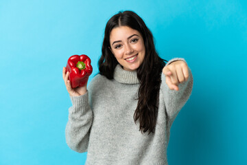 Young caucasian woman holding a pepper isolated on blue background points finger at you with a confident expression