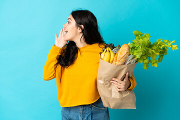 Young woman holding a grocery shopping bag isolated on blue background shouting with mouth wide open to the side