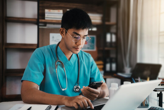 Mid Adult Male Doctor Reviews Patient Records On Smartphone And Desktop PC.