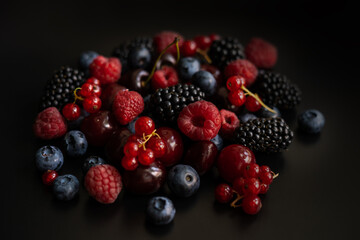 Raspberries, red currant, blueberries, blackberries and cherries are placed in a pile on a dark background 