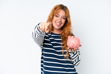 Young caucasian woman holding a piggybank isolated on white background points finger at you with a confident expression