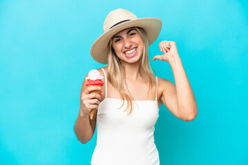 Young caucasian woman in swimsuit with a cornet ice cream isolated on blue background proud and...