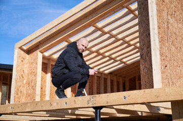 Male engineer building wooden frame house on pile foundation. Bald man standing on construction site, inspecting quality of work on sunny day.