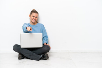 Young woman with a laptop sitting on the floor points finger at you with a confident expression