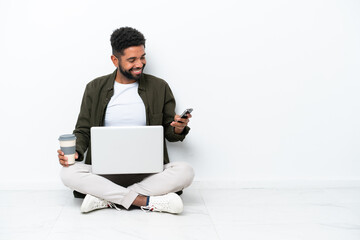 Young Brazilian man with a laptop sitting on the floor isolated on white holding coffee to take away and a mobile