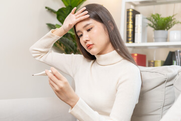 Sick, influenza asian young woman, girl headache have a fever, flu and check thermometer measure body temperature, feel illness sitting on sofa bed at home. Health care person on virus, covid-19.