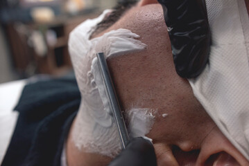 A barber uses a straight edge razor to shave the sideburns and beard of a client. Removing the...
