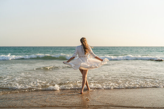 Chica joven en bata blanca de verano entrando en el mar al atardecer 