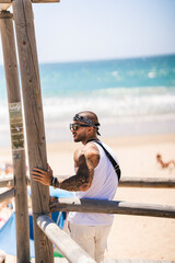 Chico joven guapo musculado y tatuado en la playa en día soleado con camiseta de tirantes blanca y...