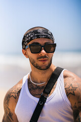 Chico joven guapo musculado y tatuado en la playa en día soleado con camiseta de tirantes blanca y pañuelo en la cabeza
