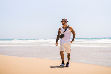 Chico joven guapo musculado y tatuado en la playa en día soleado con camiseta de tirantes blanca y...