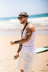 Chico joven tatuado musculoso en la playa con camiseta de tirantas blanca y pañuelo en la cabeza