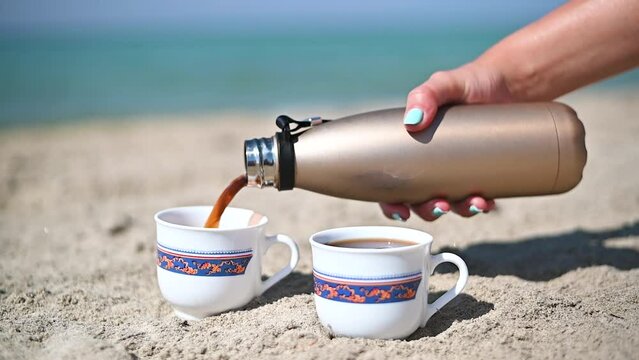 Pouring black coffee in cups on the sand beach from coffee thermostat. Slow motion