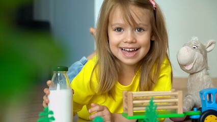 Portrait of little girl with milk bottle looks at camera and laughs. Happy child drinks milk