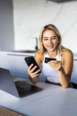 Happy young woman with laptop using credit card and phone in kitchen at home