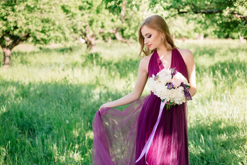 A bridesmaid in a green summer garden poses in a purple dress with a bouquet of light peonies and...