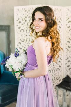 Pretty young girl in a purple dress with curly hair looks into the camera, smiled and holds a bouquet of peonies in her hands. Bridesmaid in Provencal style at home. High quality photo.