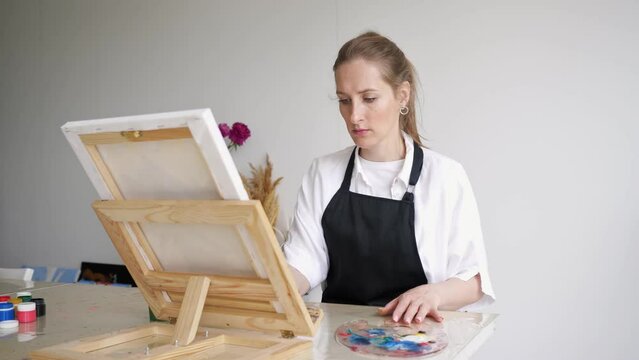 Mature woman paints picture on easel sitting at table. Professional artist enjoys painting masterpieces with paintbrush and palette closeup
