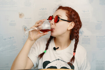 Cheerful female in round red goggles with funny pigtails drinks wine. Hippie woman holding glass of alcoholic drink. Copy space for design and conceptual blogger advertising.