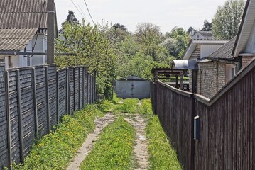 rural alley with a road in green grass along fences made of gray concrete and brown wooden boards and gray metal gates in a dead end