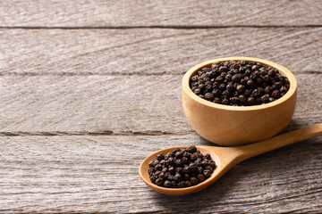 Closeup black pepper seeds or peppercorns (dried seeds of piper nigrum) in wooden bowl and spoon isolated on rustic wood table background.