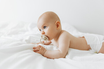 newborn baby sucks on a pacifier and lies on a white bed at home in a white sunny bedroom